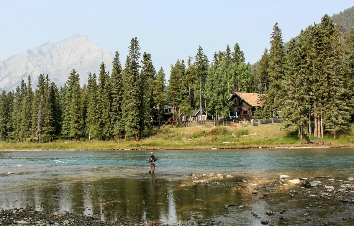 An angler fly fishing in a river beside forest cabins and distant mountains; illustrative activity photograph.